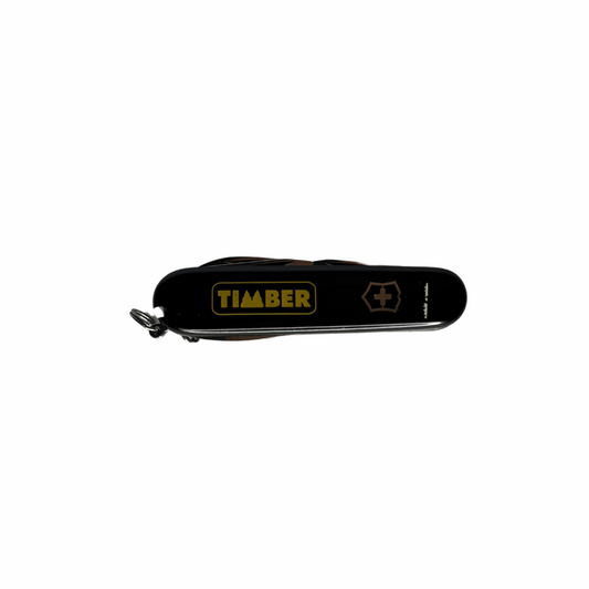 Agora fechado, com o corpo compacto em preto brilhante. A lateral apresenta o logo "TIMBER" em amarelo e o emblema da Suiça.