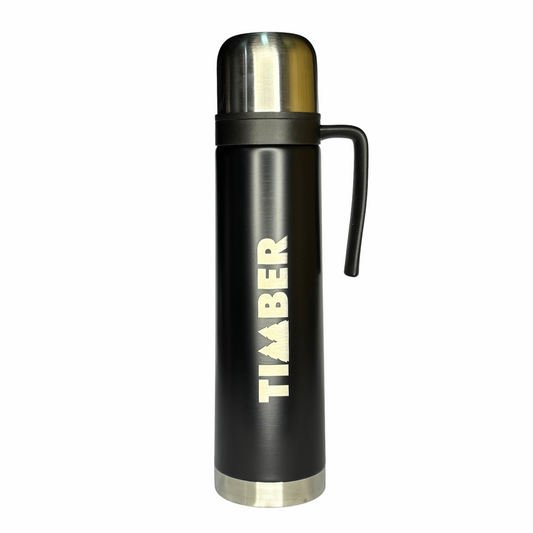 A garrafa térmica possui corpo cilíndrico preto com detalhes em aço inoxidável na base e na tampa. O logotipo "TIMBER" está em destaque na lateral, em cor branca. 