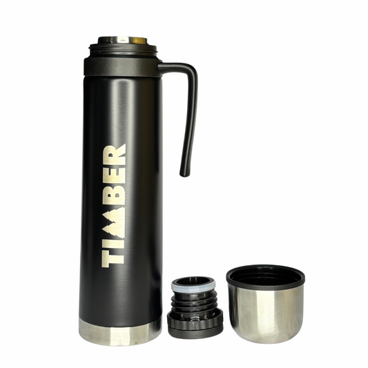 A garrafa térmica possui corpo cilíndrico preto com detalhes em aço inoxidável na base e na tampa. O logotipo "TIMBER" está em destaque na lateral, em cor branca. A tampa superior também serve como copo, enquanto uma alça preta lateral facilita o manuseio.