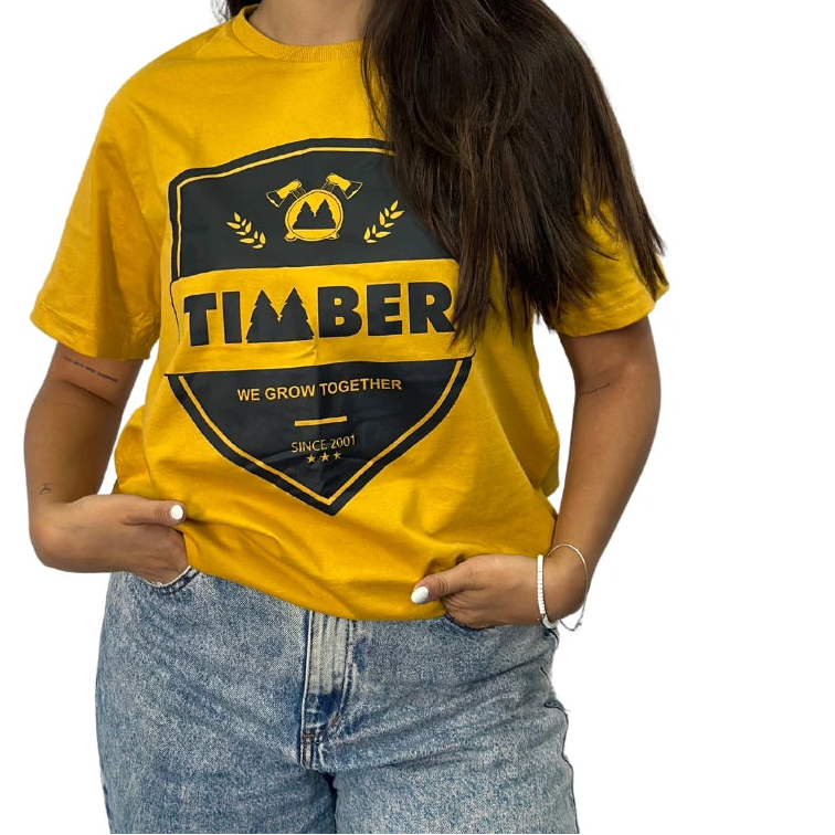 Modelo usando camiseta amarela feminina 100% algodão com estampa preta 'TIMBER' e slogan 'We Grow Together'.