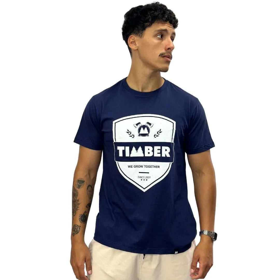 Modelo usando camiseta azul masculina 100% algodão com estampa branca 'TIMBER' e slogan 'We Grow Together'.